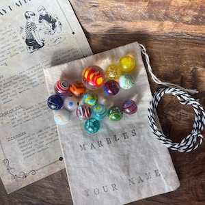 May include: A collection of colorful glass marbles in a drawstring bag with the word "MARBLES" printed on it. A vintage-style instruction sheet and a black and white rope are also included. The bag is personalized with the text "YOUR NAME".