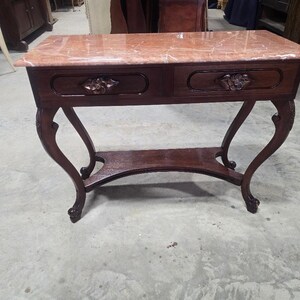 May include: A rectangular console table featuring a polished pink marble top and a dark brown wooden base. The table includes two drawers with carved floral accents on the front panels, supported by four curved legs connected by a lower shelf.