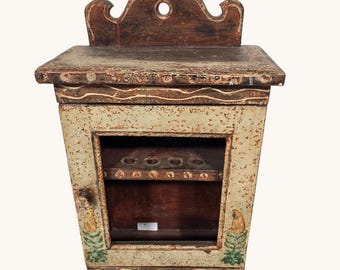 May include: A vintage wooden wall cabinet with a distressed, off-white finish. The cabinet features a decorative top, a small window, and painted floral accents. The interior has a shelf with several small holes. The cabinet is likely used for storage.