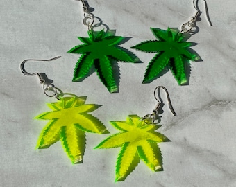 May include: A set of four earrings featuring green and yellow marijuana leaf designs. The earrings are made of acrylic and have silver hooks.