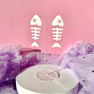 Pode incluir: Brincos de espinha de peixe branco sujo com um 'X' vermelho na cabeça do peixe. Os brincos estão pendurados em um suporte transparente contra um fundo rosa. Um acessório divertido e peculiar.