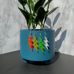 May include: Four pairs of lightning bolt earrings in vibrant colors: orange, yellow, green, and white. Each earring has a silver hook. The earrings are displayed in front of a blue plant pot with a green plant.