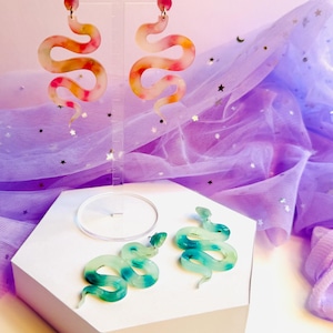 May include: Snake-shaped earrings in two styles. One pair is a long, dangling design with a pink, yellow, and orange pattern. The other pair is a smaller, stud-style earring in shades of green and blue. Both sets are displayed on a white hexagonal stand.
