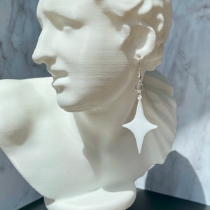 May include: White star-shaped dangle earrings with a silver hook. The earrings are hanging from a white mannequin's ear.