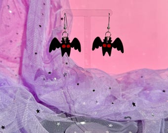May include: A pair of dangle earrings featuring black bat-shaped charms with bright red circular eyes. The earrings are suspended from silver-toned metal hooks and are displayed on a clear acrylic stand against a pink and purple backdrop.