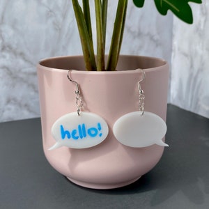 May include: A pair of white speech bubble earrings. One earring has the word "hello!" written in blue. The earrings are hanging from a plant pot.