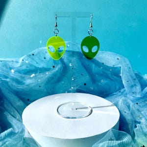 May include: A pair of alien head earrings. Each earring features a green, oval-shaped alien head with large, oval eye cutouts. The earrings hang from silver-colored hooks. The background is a light blue with a white circular stand.