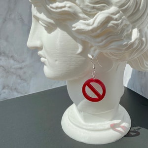 Puede incluir: Pendientes de círculo rojo con una línea diagonal que atraviesa el centro, colgando de una cadena plateada. Los pendientes los lleva un busto de yeso blanco.