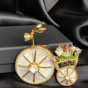 May include: A gold-coloured bicycle brooch with white and gold wheels, a basket of flowers, and a decorative headlight. The brooch features an iridescent gem and is set against a black background.