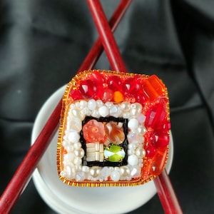 Può includere: Spilla artigianale a forma di rotolo di sushi, con perline e gemme rosse, arancioni, bianche e verdi. La spilla è impreziosita da filo color oro e poggia su un piatto bianco, con bacchette rosse incrociate dietro.