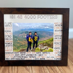 May include: A framed photograph with the text "NH 48 4000 FOOTERS" at the top. The image features two people holding gold balloons on a mountain peak. The frame is dark brown, and the names of the mountains are listed around the photo.