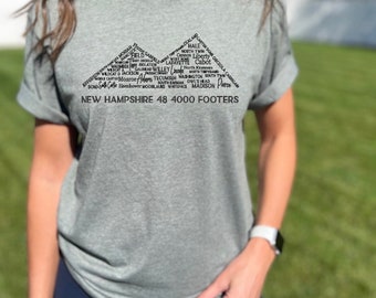May include: A heather grey t-shirt with a black graphic design of the names of 48 mountains in New Hampshire, arranged in the shape of two mountain peaks. The text "NEW HAMPSHIRE 48 4000 FOOTERS" is printed below the graphic.