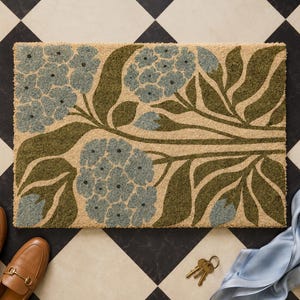 May include: A rectangular coir doormat featuring a botanical print with large, light blue hydrangea flower clusters and dark olive green leaves set against a natural tan background. The design covers the entire surface of the mat.