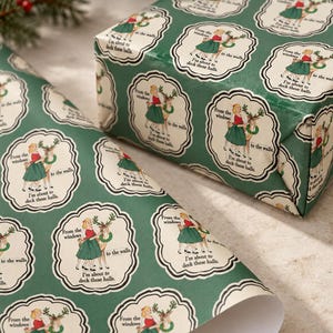 May include: Green wrapping paper featuring a repeating pattern of cream-coloured scalloped frames. Each frame contains an illustration of a person in a red top and green skirt with a reindeer, and the text: From the windows to the walls, I'm about to deck these halls.