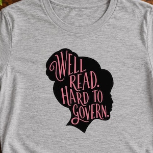 Puede incluir: Una camiseta gris jaspeado de manga corta con una silueta negra de perfil de una cabeza. Dentro de la silueta, el texto rosa dice: Well Read. Hard to Govern. La camiseta tiene un cuello redondo y un detalle de nudo en el dobladillo.