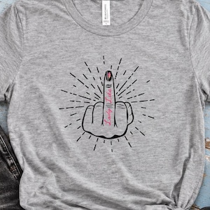Puede incluir: Una camiseta gris jaspeado de manga corta con un gráfico negro de una mano haciendo el gesto del dedo medio, con el texto "Lady Like" escrito en rosa sobre el dedo, rodeado de líneas negras radiantes.