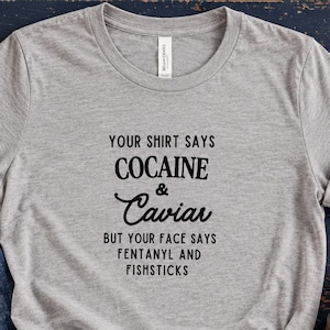 Puede incluir: Una camiseta gris jaspeado de manga corta con texto negro que dice: YOUR SHIRT SAYS COCAINE & Caviar BUT YOUR FACE SAYS FENTANYL AND FISHSTICKS. La camiseta se muestra junto a unos vaqueros azules, botines negros y un collar dorado.