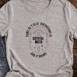 Puede incluir: Una camiseta gris jaspeado de manga corta con un estampado gráfico negro que dice "SOME OF Y'ALLS BIRTHSTONE IS CRYSTAL METH AND IT SHOWS", centrado en un marco ovalado que contiene un ojo, un cristal y símbolos celestiales.