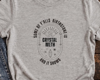 Op de afbeelding: Een grijs gemêleerd T-shirt met korte mouwen en een zwarte grafische print met de tekst "SOME OF Y'ALLS BIRTHSTONE IS CRYSTAL METH AND IT SHOWS", gecentreerd in een ovaal kader met een oog, een kristal en hemelse symbolen.