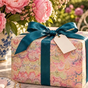 Puede incluir: Una caja de regalo rectangular envuelta en papel con un patrón de mármol multicolor en tonos rosa, amarillo y azul. La caja está atada con un lazo de cinta de satén de color verde azulado oscuro y tiene una etiqueta de regalo color crema.