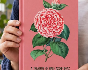 Op de afbeelding: Een roze notitieboek met harde kaft voorzien van een gedetailleerde illustratie van een bloeiende camelia met groene bladeren en de tekst A TREASURY OF HALF ASSED IDEAS in het zwart onderaan het midden.