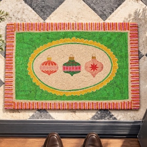 May include: A rectangular doormat featuring a bright green border with pink and orange stripes. The center has a beige oval framed in yellow, displaying three colorful Christmas ornaments in red, green, and pink with star and stripe patterns.