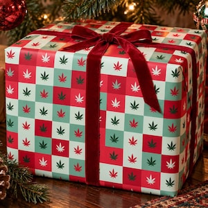 May include: A rectangular gift box wrapped in paper featuring a chequered pattern of red, light green, and dark green squares, each decorated with a small white cannabis leaf icon. The box is tied with a dark red velvet ribbon and bow.