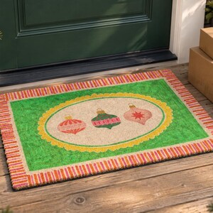 May include: A rectangular doormat featuring a bright green center with a central oval containing three illustrated Christmas ornaments in red, green, and pink. The mat has a decorative border with alternating red and orange vertical stripes.