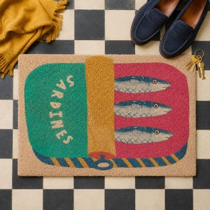 May include: A rectangular doormat featuring a graphic of an open sardine tin. The design includes a green section with the text SARDINES, a yellow lid, and a pink section containing three blue fish illustrations with a blue and yellow striped border.