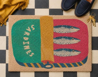 May include: A rectangular doormat featuring a graphic of an open sardine tin. The design includes a green section with the text SARDINES, a yellow lid, and a pink section containing three blue fish illustrations with a blue and yellow striped border.