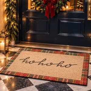 Puede incluir: Un felpudo rectangular de fibra de coco color canela con un borde de cuadros rojos, verdes y negros. El centro de la alfombra muestra el texto "hohoho" escrito en una fuente cursiva negra. La alfombra est&aacute; colocada sobre un suelo de cuadros.