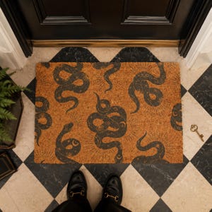 May include: A rectangular coir doormat featuring a repeating pattern of dark gray snakes intertwined with celestial motifs, including sunbursts, stars, and crescent moons, printed against a natural tan fibrous background.