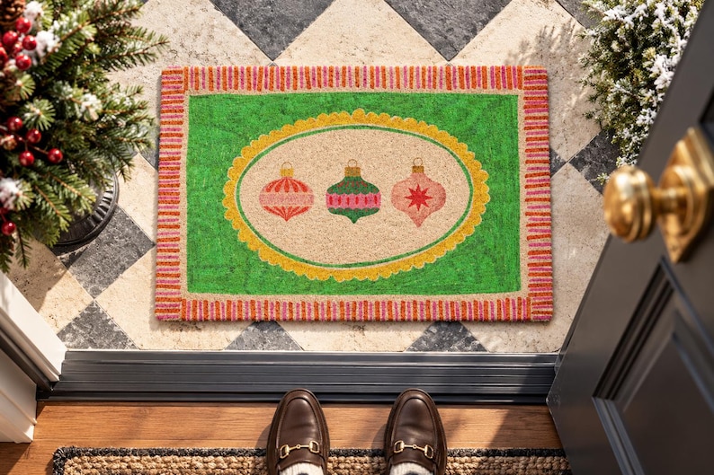 May include: A rectangular doormat featuring a bright green border with pink and orange stripes. The center has a beige oval framed in yellow, displaying three colorful Christmas ornaments in red, green, and pink with star and stripe patterns.