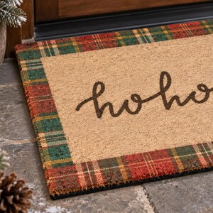 Puede incluir: Un felpudo rectangular de fibra de coco color canela con el texto hohoho en letra cursiva de color marr&oacute;n oscuro. La alfombra est&aacute; bordeada por un patr&oacute;n de cuadros festivo en rojo, verde y dorado. La textura es rugosa y fibrosa.