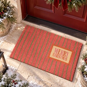 May include: A rectangular coir doormat featuring a red background with thin vertical green stripes. A central rectangular tan patch displays the text Merry Christmas in red lettering. The mat has a coarse, textured surface typical of natural fiber.