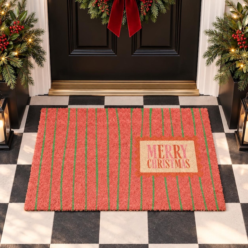 May include: A rectangular doormat with a coral red background and thin vertical green stripes. A central rectangular tan patch features the text Merry Christmas in pink and red lettering. The mat has a textured, fibrous surface appearance.