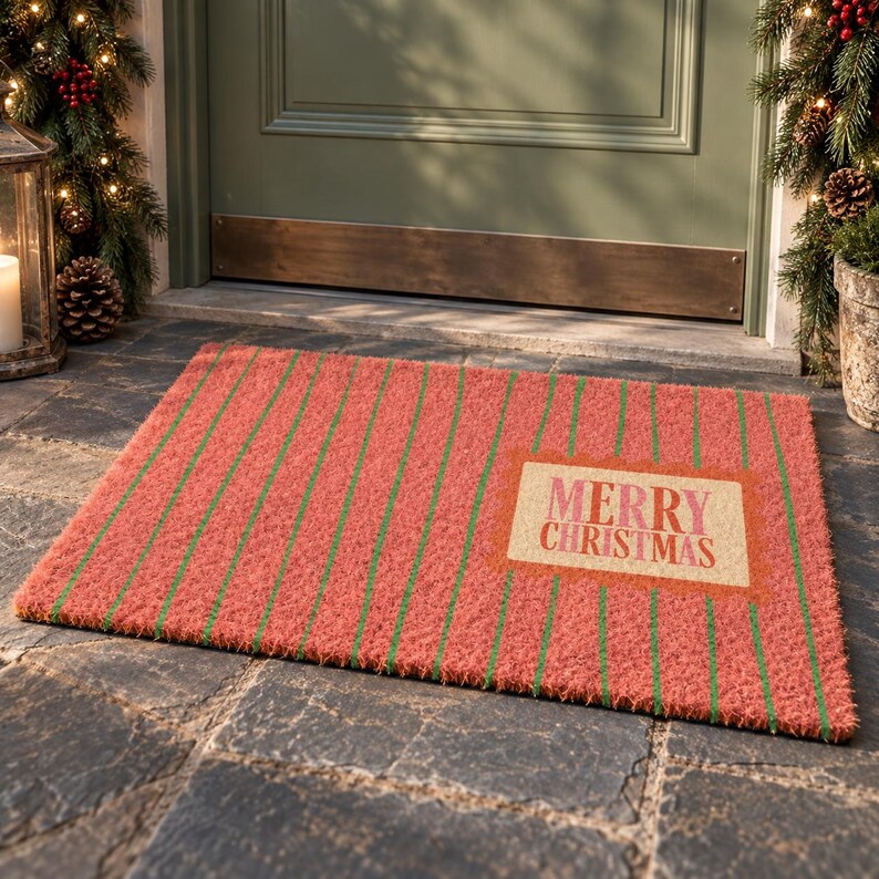 May include: A rectangular doormat with a textured surface featuring vertical red and green stripes. A central rectangular cream-colored patch displays the text MERRY CHRISTMAS in red and pink lettering. The mat has a coarse, fibrous appearance.