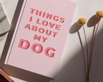 Op de afbeelding: Een roze notitieboek met de tekst "THINGS I LOVE ABOUT MY DOG" in een gelaagde, rode letter. Het gesloten notitieboek ligt op een wit oppervlak, met twee gele gedroogde bloemen rechts.