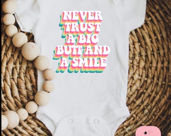 Op de afbeelding: Witte babybody met de tekst "NEVER TRUST A BIG BUTT AND A SMILE" in een retro, kleurrijk lettertype. De tekst is gelaagd met roze, oranje, geel, groen en blauw. De body heeft drukknoopsluitingen en korte mouwen.