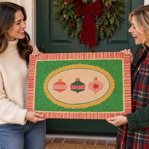 May include: A rectangular coir doormat featuring a green border with a striped red and orange edge. The center has a tan oval with a scalloped yellow frame, displaying three illustrated Christmas ornaments in shades of red, green, and pink.