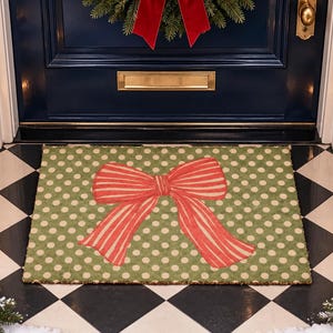 May include: A rectangular doormat featuring a sage green background with a repeating pattern of small cream-coloured polka dots. Centered on the mat is a large, stylised red bow with thin vertical stripes, creating a festive and seasonal appearance.