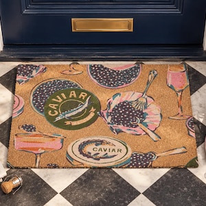 May include: A rectangular coir doormat featuring a repeating pattern of caviar tins, champagne glasses, and serving spoons. The design includes circular green and tan tins labeled CAVIAR, pink champagne coupes, and flutes on a natural brown background.