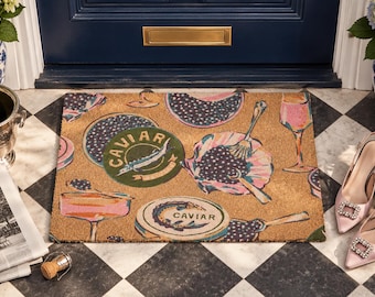 May include: A rectangular coir doormat featuring a repeating pattern of caviar tins, champagne glasses, and serving spoons. The design includes circular green and tan tins labeled CAVIAR, pink champagne coupes, and flutes on a natural brown background.