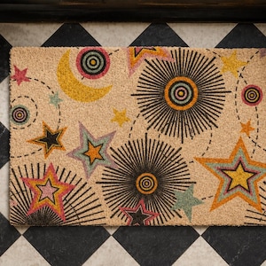 May include: A rectangular coir doormat featuring a celestial pattern with a yellow crescent moon, various multi-colored stars, and black sunburst motifs. The design includes dashed circular lines connecting the shapes across the tan surface.