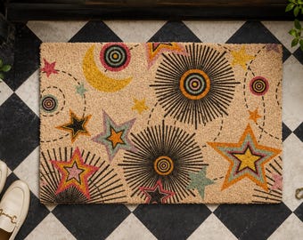 May include: A rectangular coir doormat featuring a celestial pattern with a yellow crescent moon, various multi-colored stars, and black sunburst motifs. The design includes dashed circular lines connecting the shapes across the tan surface.