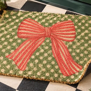 May include: A rectangular coir doormat featuring a green background with a pattern of light tan polka dots. Centered on the mat is a large, prominent red bow with thin, vertical white stripes, creating a festive and decorative appearance for an entryway.