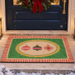 May include: A rectangular doormat featuring a central beige oval with three decorative Christmas ornaments in red, green, and pink. The mat has a green border surrounded by a red and gold striped frame, set against a snowy outdoor entryway.