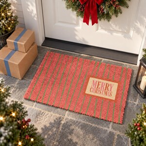 May include: A rectangular coir doormat featuring a red background with thin vertical green stripes. A centered rectangular tan label displays the text Merry Christmas in red and pink lettering. The mat has a coarse, textured surface finish.