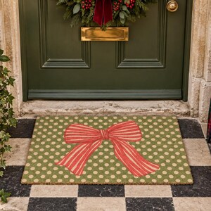 May include: A rectangular doormat featuring a repeating pattern of light beige polka dots on a muted olive green background, centred with a large, prominent red bow with thin vertical stripes, positioned on a black and white chequered floor.