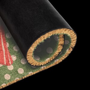 May include: A rolled doormat with a black rubber backing and a natural coir fibre front. The visible surface features a green background with a pattern of light-coloured polka dots and a portion of a red graphic design on the left side.